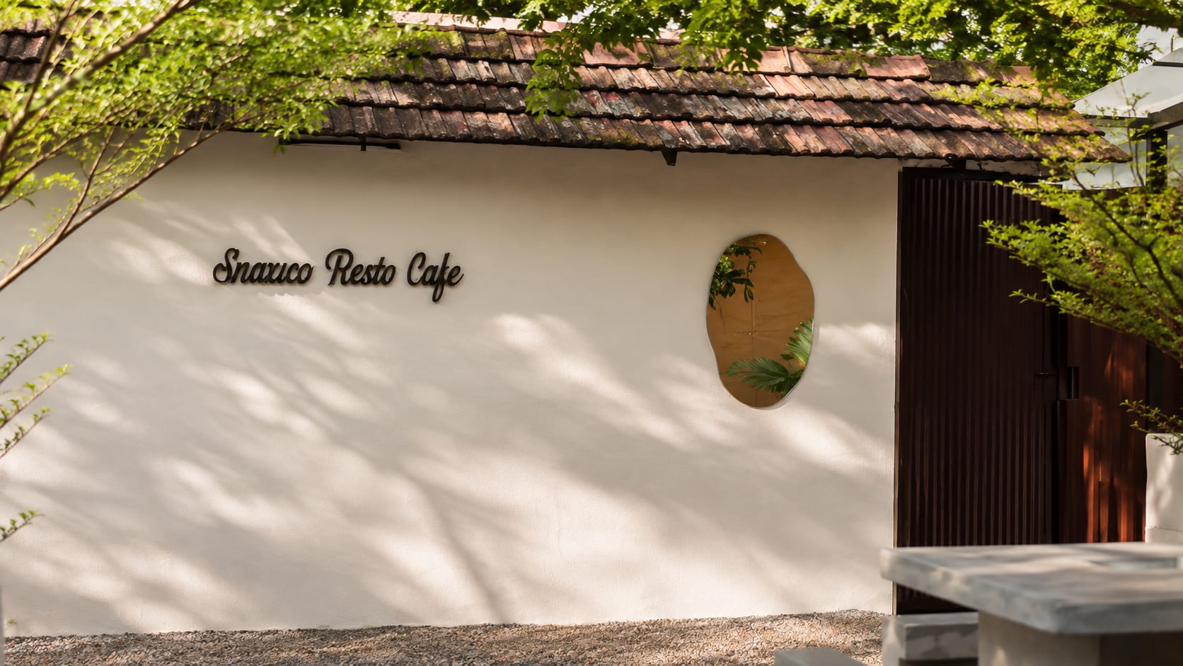 Snaxico Resto Cafe white stucco wall with script wordmark, clay tile roof, and organic cutout window