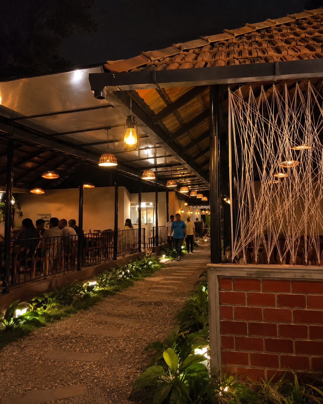 Lantern-lit walkway under clay tile roof with string art and dining tables at night